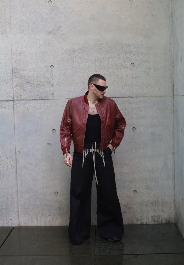 Uomo con occhiali da sole neri, giacca di pelle rossa, canotta nera e pantaloni neri larghi in piedi contro un muro di cemento.