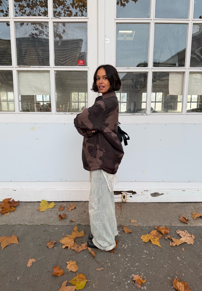 Camo-patterned oversized jacket in brown tones, paired with light linen wide-leg pants. Black boots on a leaf-strewn pavement.