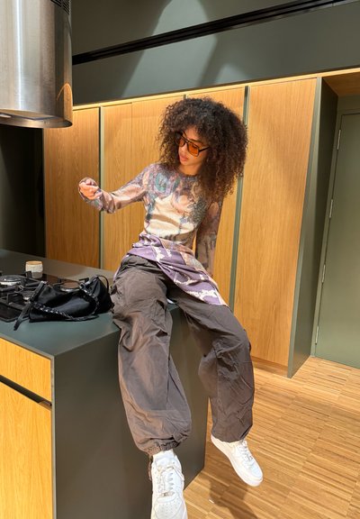 Blusa de manga larga con un estampado gráfico, combinada con pantalones oscuros sueltos y zapatillas blancas. Los accesorios incluyen gafas de sol de gran tamaño y un bolso negro.