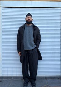 Abrigo exterior negro, suéter gris con textura, pantalones de pierna ancha a rayas blancas y negras, y zapatos negros; de pie frente a un fondo con persianas cerradas.