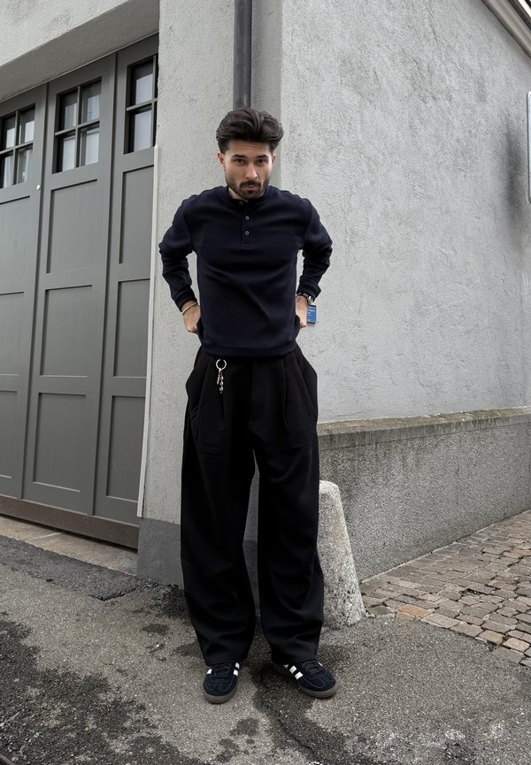 Moški v črni srajci z dolgimi rokavi in širokih črnih hlačah stoji na vogalu ulice ob sivi stavbi z garažnimi vrati v bližini.