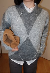 Pull gris à motif avec un col en V, présentant un design en diamant et un tricot texturé. Tenant un sac plié en suède brun. Chemise blanche en dessous.