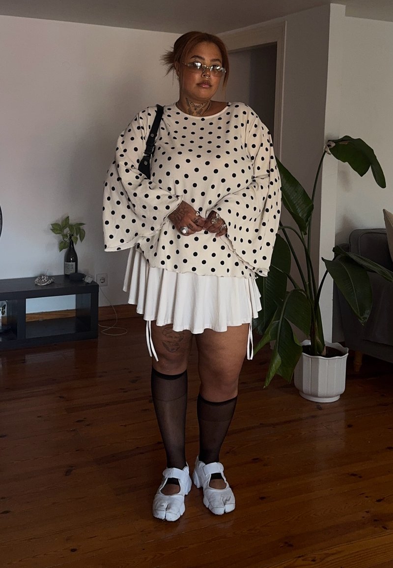 Personne portant un haut blanc à pois, une jupe plissée blanche, des chaussettes noires transparentes jusqu’aux genoux et des chaussures blanches à bout fendu, debout à l’intérieur.