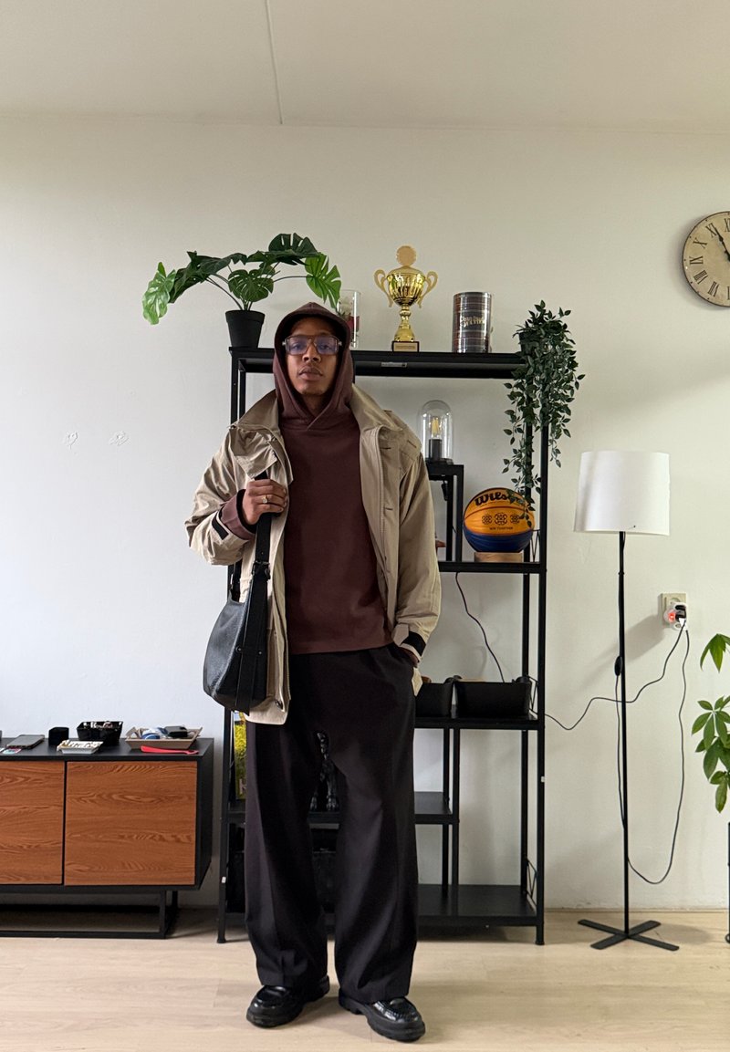 Personne portant une veste beige et un sweat à capuche marron tenant un sac noir, debout à l'intérieur près d'une étagère noire avec des plantes, des trophées et un ballon de basket.