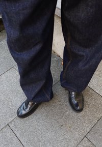 Dunkelblaue Jeans mit sichtbaren Nähten, kombiniert mit glänzenden schwarzen Slippern. Der Hintergrund zeigt grauen Steinpflaster.