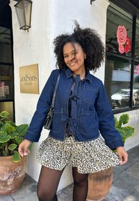 Jeansjacke mit Bindegürtel und vorderen Taschen, kombiniert mit einem gerafften Rock im Leopardenmuster. Lockige Haare, schwarze Strumpfhose und eine kleine Umhängetasche.