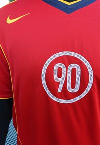 Sarkans sporta krekls ar apaļu grafiku, kurā ir "90" baltā perforētā tekstūrā. Dzeltenas un tumši zila akcenti uz apkakles. Nike logo uz pleca.