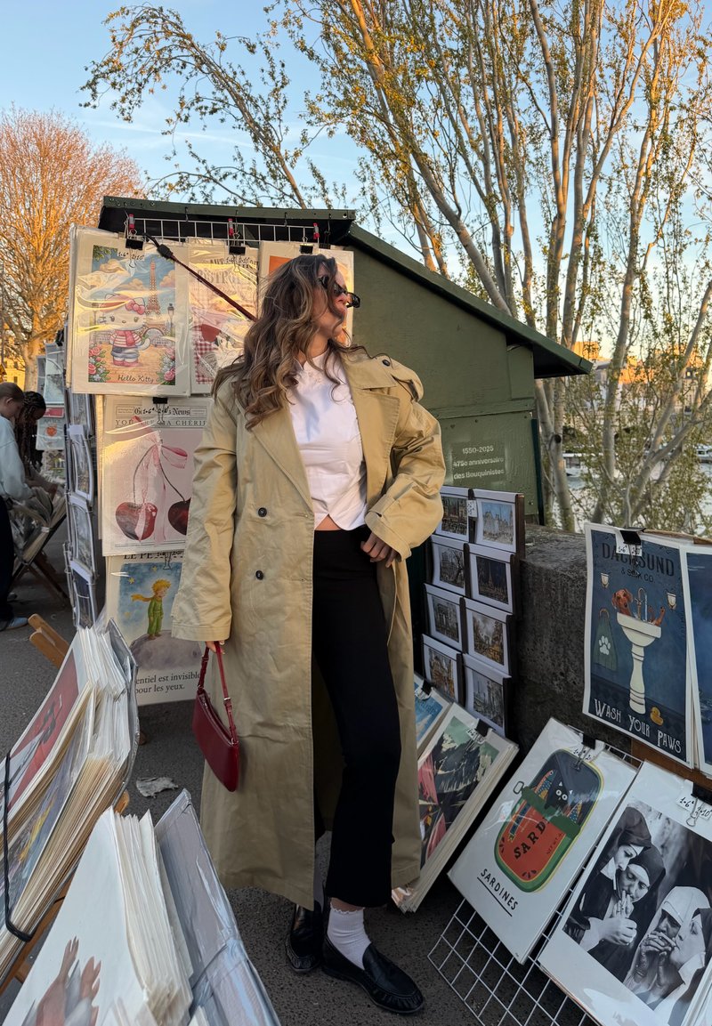 Femme en trench beige et lunettes de soleil posant dans un marché d'art en plein air avec des impressions exposées sur des présentoirs et stands à la lumière du tard après-midi.