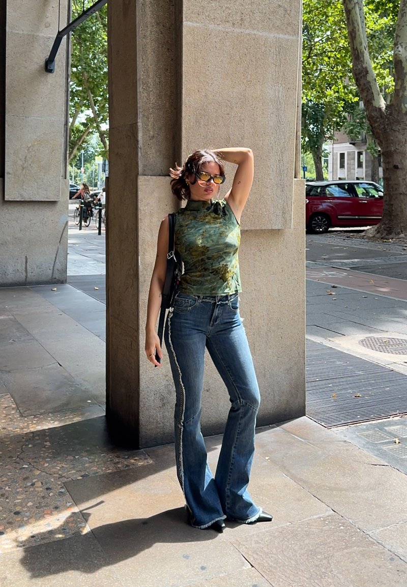 Jeune femme portant des lunettes de soleil et un haut vert sans manches pose contre un pilier en pierre sur un trottoir urbain ensoleillé.