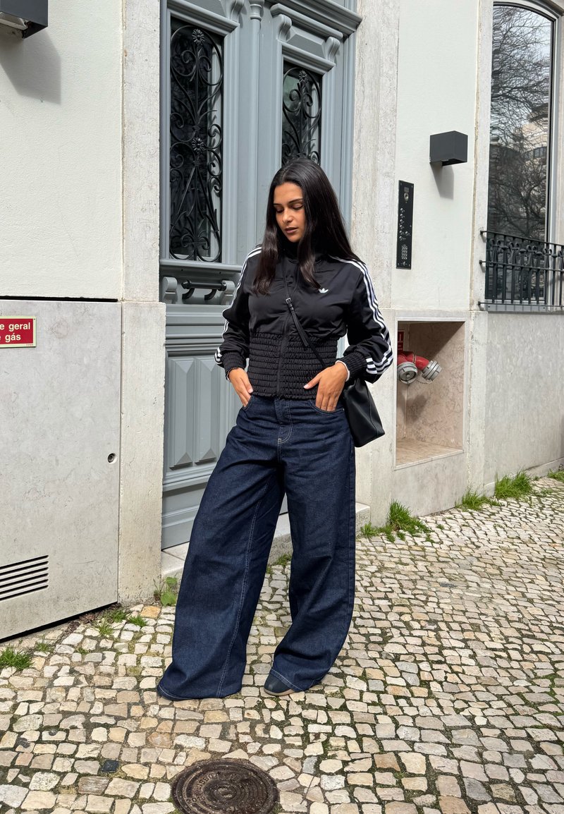 Γυναίκα με μακριά σκούρα μαλλιά, ντυμένη με μαύρο μπουφάν με λευκές ρίγες και φαρδιά σκούρα μπλε τζιν, στέκεται σε λιθόστρωτο δρόμο κοντά σε μια γκρίζα πόρτα.