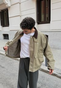 Veste vert olive avec un col en velours côtelé marron, des poches avant, et un intérieur à carreaux, portée sur un t-shirt blanc et un pantalon gris plissé.