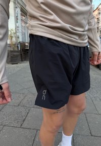 Személy fekete sportnadrágot és bézs színű hosszú ujjú felsőt visel, városi járdán áll, a combján látható tetoválással.