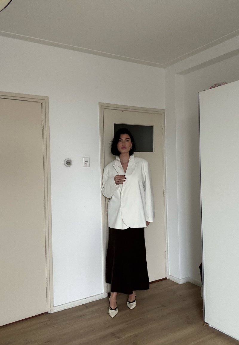Femme aux cheveux courts et foncés portant un blazer blanc oversize, une longue jupe noire et des talons blancs pointus, debout dans une pièce minimaliste.