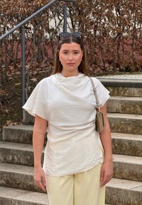 Λευκή υφασμάτινη μπλούζα με λαιμόκοψη cowl, σε συνδυασμό με ανοιχτό κίτρινο παντελόνι φαρδιάς γραμμής. Συνοδευμένη από μια μικρή τσάντα και γυαλιά ηλίου.