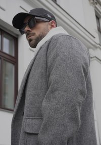 Casaco de lã cinza com colarinho em bico e bolsos frontais, sobreposto a um lenço branco, combinado com um chapéu preto e óculos de sol.