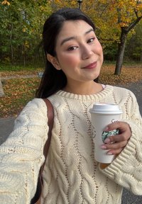 Creme kabelgebreide trui met lange mouwen, houdt een witte mok met het Starbucks-logo vast terwijl hij buiten tussen de herfstbladeren staat.