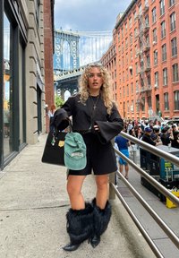 Svart cropped jakke med klokkeermer, sorte shorts, lodne sorte støvler, grønn caps med "BROOKLYN" tekst, holder en svart håndveske. Urban setting.