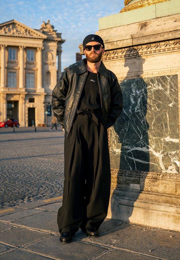 Man die een zwarte zonnebril, een leren jas en wijde zwarte broek draagt, staat bij een stenen monument op een zonnig stedelijk plein.