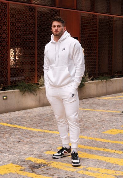 Sudadera blanca de Nike con bolsillo delantero y logo, combinada con pantalones deportivos blancos a juego. Zapatillas Nike en blanco y negro con acentos rojos.