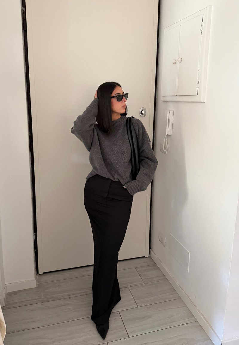 Grauer Strickpullover, schwarze taillierte Rock und Sonnenbrille; elegantes Design mit einer strukturierten Stoffoberfläche. Im Hintergrund ist eine hellfarbige Tür zu sehen.