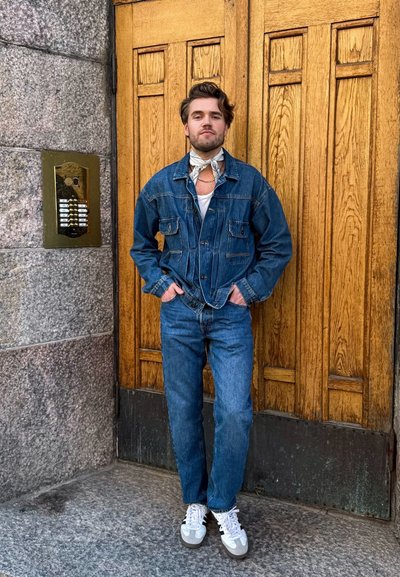 Chaqueta de mezclilla con bolsillos de parche, jeans de color azul claro, camiseta blanca y bufanda con estampado; enmarcados por puertas de madera y una pared de piedra.