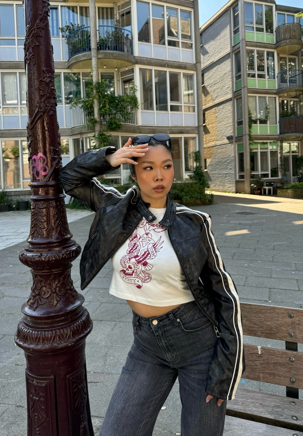 Veste en cuir noire courte avec des rayures blanches, t-shirt graphique blanc avec un design de tête de mort, jeans en denim bleu, cadre urbain avec des bâtiments.