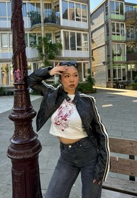 Veste en cuir noire courte avec des rayures blanches, t-shirt graphique blanc avec un design de tête de mort, jeans en denim bleu, cadre urbain avec des bâtiments.