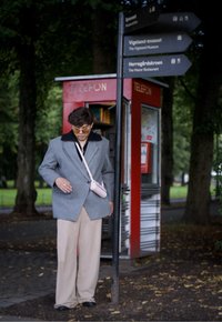 Γκρι υπερμεγέθη σακάκι με μαύρο γιακά, μπεζ παντελόνι σε φαρδιά γραμμή και ροζ τσάντα ώμου. Το φόντο περιλαμβάνει μία κόκκινη τηλεφωνική θαλάμου και κατευθυντήριες πινακίδες.