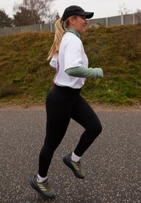 Tenue de sport composée d'un T-shirt blanc, de manches longues vertes en dessous, de leggings noirs taille haute et de chaussures de course vert olive avec des accents noirs.