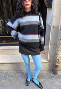 Άτομο που φοράει πουλόβερ με ρίγες και χνουδωτή υφή, μαύρη φούστα, φωτεινές μπλε κάλτσες, μαύρα παπούτσια με τετράγωνη μύτη και γυαλι ά ηλίου, στέκεται σε πεζοδρόμιο πόλης.