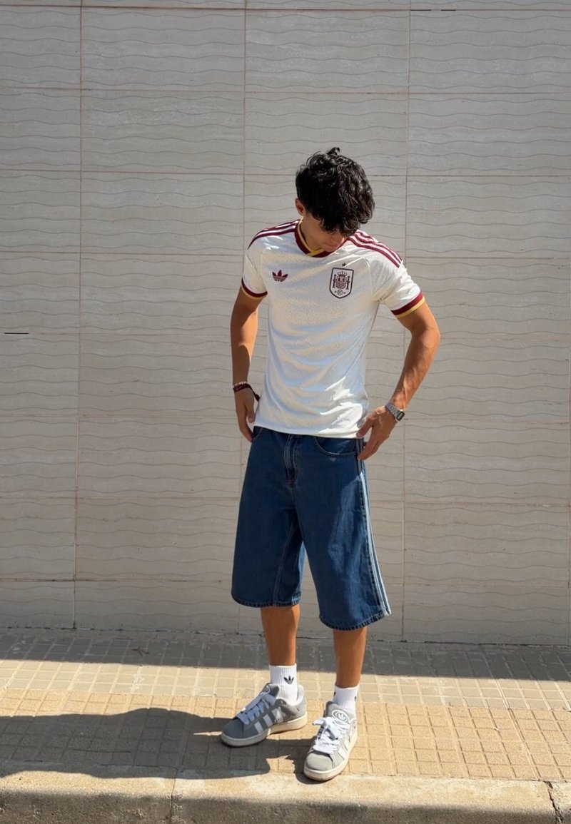 Jeune homme portant un maillot de football espagnol blanc, un long short en jean et des baskets grises, debout sur le trottoir, regardant vers le bas avec les mains près des poches.