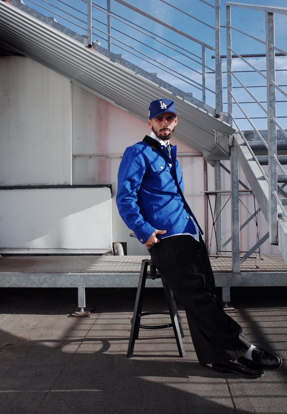 Modrá džínsová bunda s čiernymi detailmi na golieri, pod ňou biela košeľa, čierne nohavice a čierne topánky. Model sedí na čiernom stoličke.