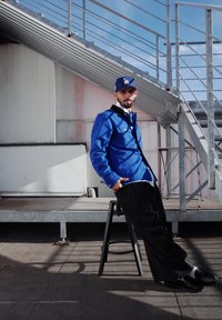 Modrá džínsová bunda s čiernymi detailmi na golieri, pod ňou biela košeľa, čierne nohavice a čierne topánky. Model sedí na čiernom stoličke.
