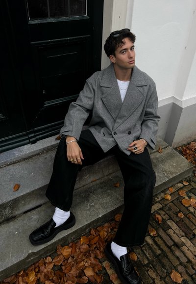 Abrigo de lana gris de doble botonadura sobre una camisa blanca, combinado con jeans negros de pierna ancha y zapatos negros sin cordones con calcetines blancos. El fondo tiene hojas caídas.