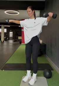Femme debout sur un tapis gris, soulevant des haltères noirs latéralement dans une salle de sport avec du gazon vert et des murs noirs.