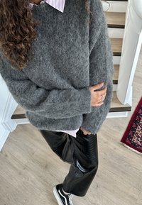 Grijze, zachte gebreide trui met lange mouwen, gedragen over een gestreept roze-wit overhemd met kraag, gecombineerd met zwarte, glanzende broek met wijde pijpen en sneakers.