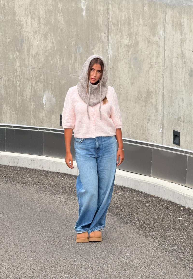 Cardigan curto de manga curta, texturizado, em rosa claro, com botões; jeans azuis de ganga folgados; cachecol cinzento de malha; sapatos baixos em tom bege; fundo neutro.