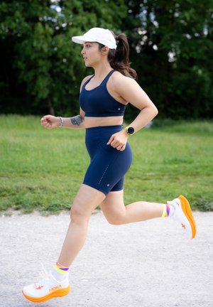 Sujetador deportivo navy y pantalones cortos; gorra blanca; zapatillas de correr blancas con suelas naranjas; calcetines coloridos; entorno al aire libre con hierba de fondo.