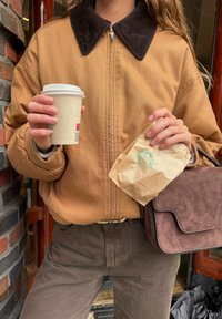 Giacca bomber beige con colletto in velluto a coste scuro, pantaloni marroni, tiene in mano una tazza di carta per caffè e una busta di carta marrone. Presenta materiali texture.