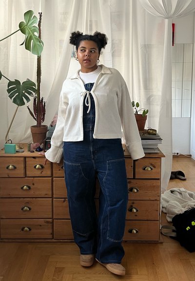 Chaqueta blanca con textura y un diseño anudado al frente, sobre un mono de mezclilla azul con piernas anchas y bolsillos. En el fondo, un tocador de madera natural.