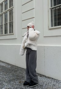 Λευκή γούνα ζακέτα με πλεκτές λεπτομέρειες, γκρι ριγέ παντελόνι και κρεμ πλεκτό σκουφάκι. Μαύρα παπούτσια με χοντρή σόλα. Στέκεται κοντά σε έναν τοίχο.