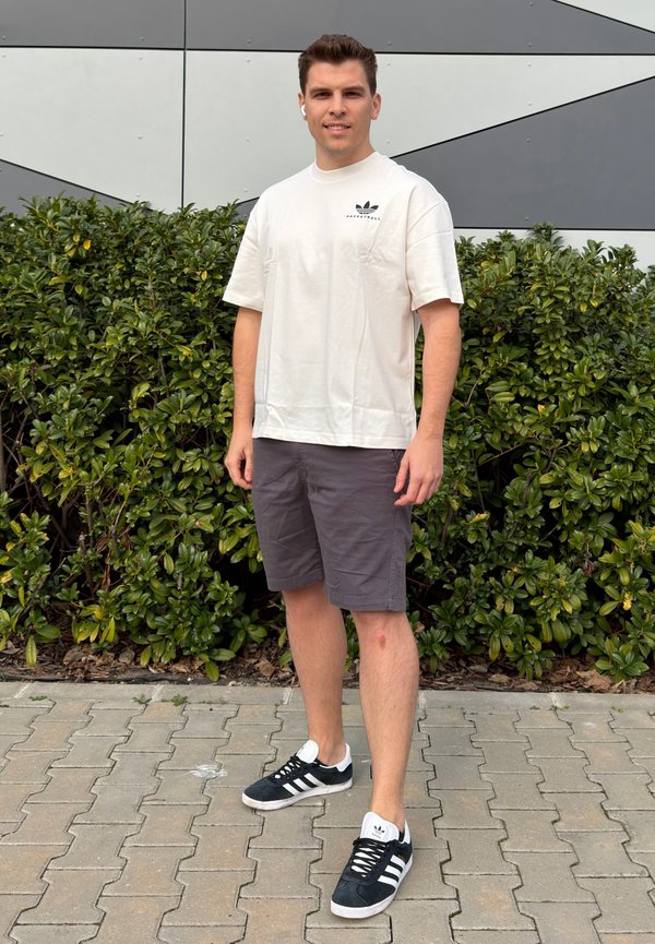 Jaunas vyras, dėvintis baltą Adidas marškinėlį, pilkus šortus ir juodus Adidas sportinius batelius, stovi ant šaligatvio priešais žalius krūmus ir geometrinę sieną.