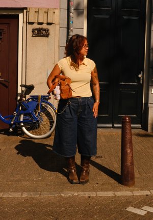 Jeans-Culottes in einer dunklen Waschung, kombiniert mit einem hellgelben Poloshirt. Braune Knöchelschuhe. Eine hellbraune Tasche mit einem Puppenanhänger. Blaues Fahrrad im Hintergrund.