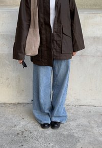 Barna, oversized dzseki elülső cipzárral és zsebbel, világoskék, bő szárú farmernadrággal és fekete cipővel párosítva. Egy bézs velúr táska lóg az egyik vállán.