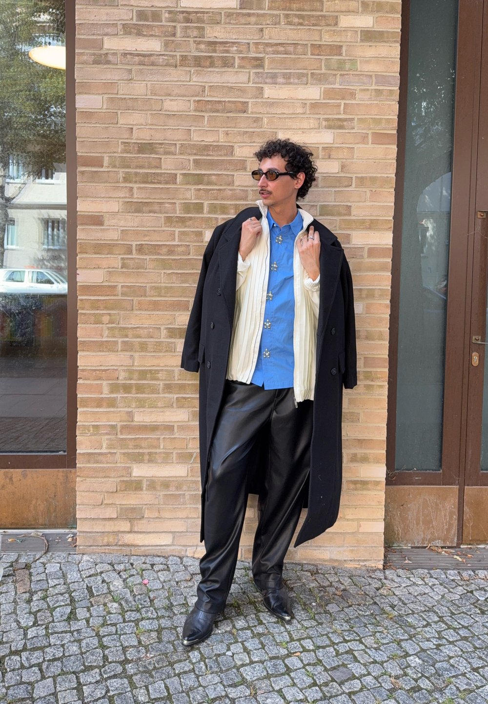Uomo con occhiali da sole, cappotto lungo nero sopra una giacca bianca e una camicia blu, pantaloni in pelle nera, in piedi su un pavimento di ciottoli vicino a un muro di mattoni.