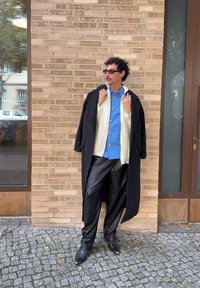 Uomo con occhiali da sole, cappotto lungo nero sopra una giacca bianca e una camicia blu, pantaloni in pelle nera, in piedi su un pavimento di ciottoli vicino a un muro di mattoni.