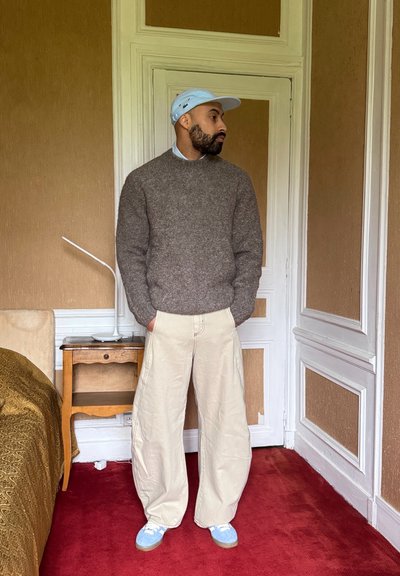 Suéter de punto de bronce, pantalones holgados beiges y zapatos azul claro. Gorra simple y una camisa con collar visibles. De pie en una habitación con una alfombra roja.