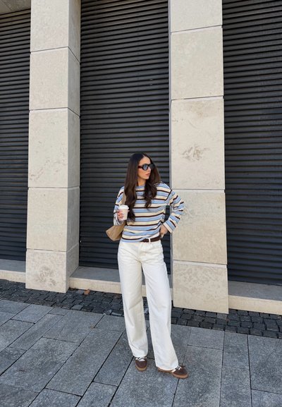 Mujer con gafas de sol, camiseta de manga larga a rayas, pantalones blancos y zapatos marrones, sosteniendo una taza de café y de pie frente a una pared de piedra y persianas negras.