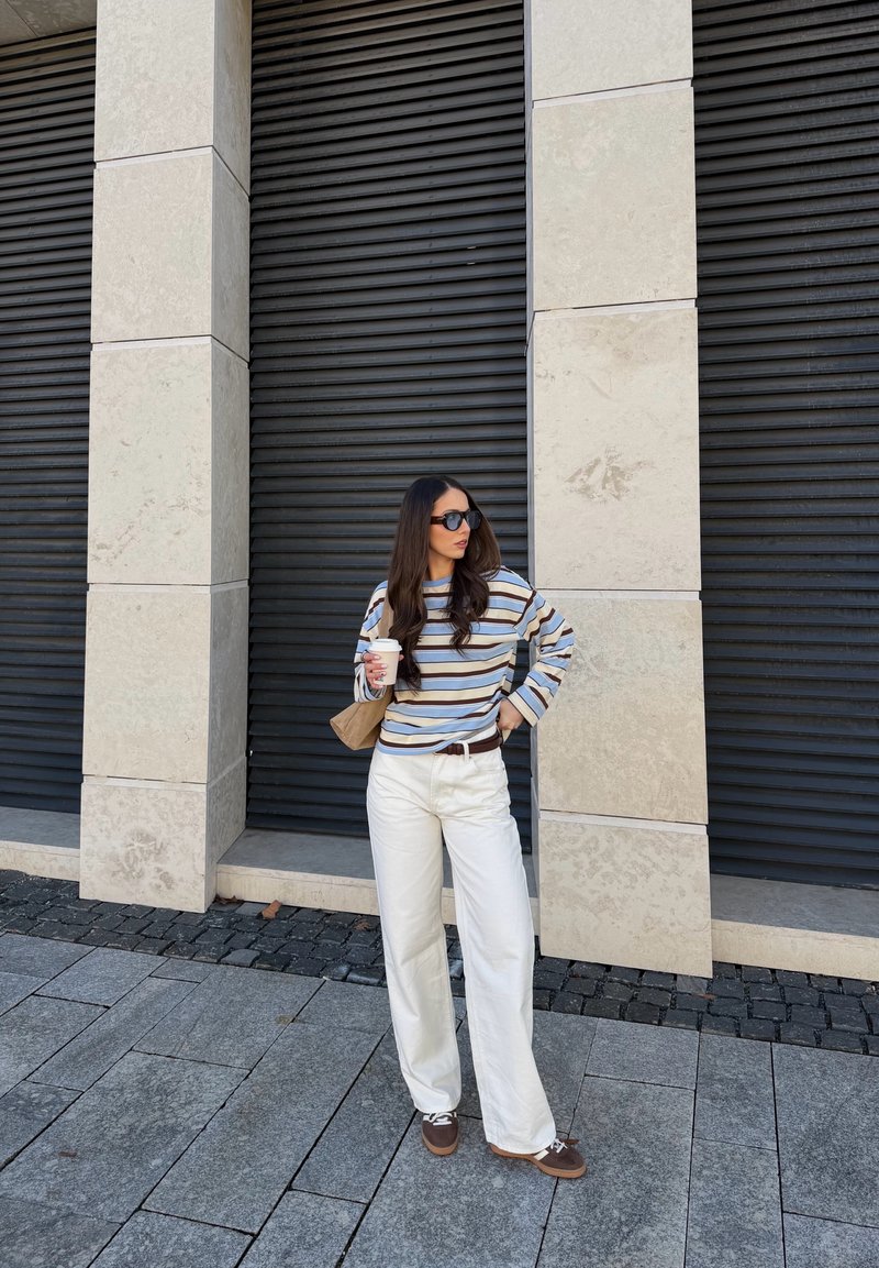 Frau mit Sonnenbrille, gestreiftem Langarmshirt, weißer Hose und braunen Schuhen, die eine Kaffeetasse hält und vor einer Mauer aus Stein und schwarzen Fensterläden steht.