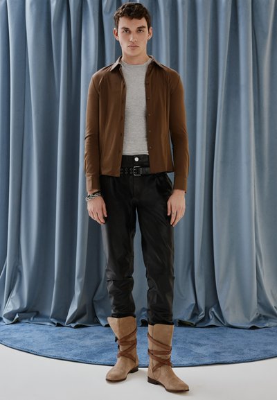 Camisa de botones marrón sobre una camiseta gris, pantalones de cuero negros y botas de color arena con tiras marrones. De pie frente a un fondo azul.